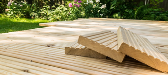 Terrassenbau mit hellen Lärchenholzdielen, gestapelte Riffelbretter im Vordergrund, umgeben von grünem Garten.