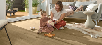 Helles Parkett im Wohnzimmer. Eine Frau spielt mit einem kleinen Mädchen auf dem Boden.